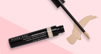 An open tube of Mary Kay® Perfecting Concealer is photographed alongside its doe-foot applicator and a product smear in front of a two-toned pink background.