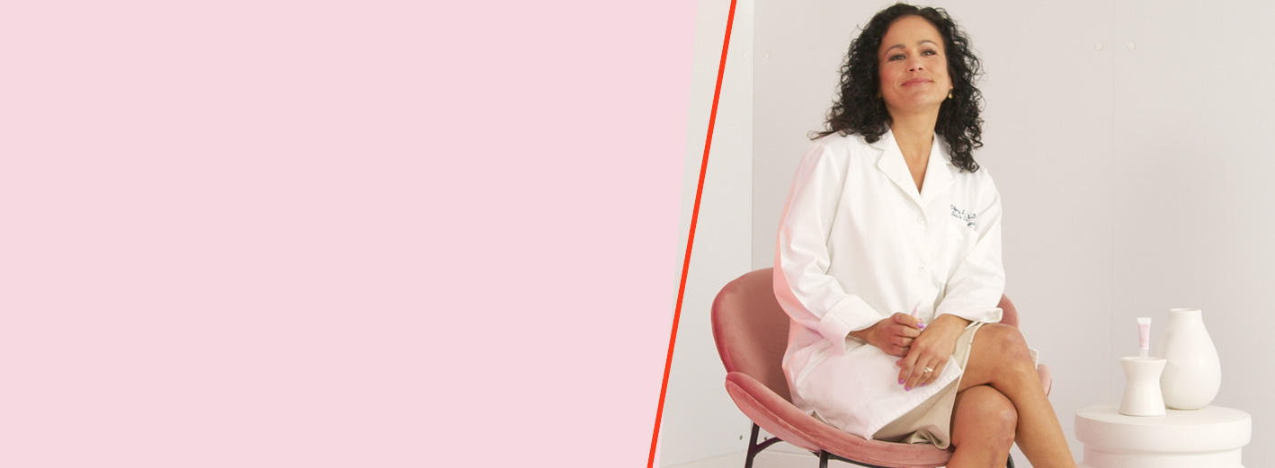Científica de Mary Kay sentada en una silla rosa junto a una mesa con un tubo de Mary Kay® Instant Puffiness Reducer sobre un fondo blanco