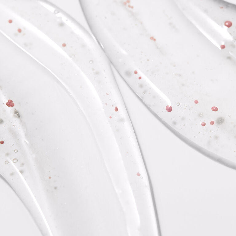 Close-up of clear facial serum spread in smooth, curved streaks with small pink beads suspended throughout, highlighting the product texture.