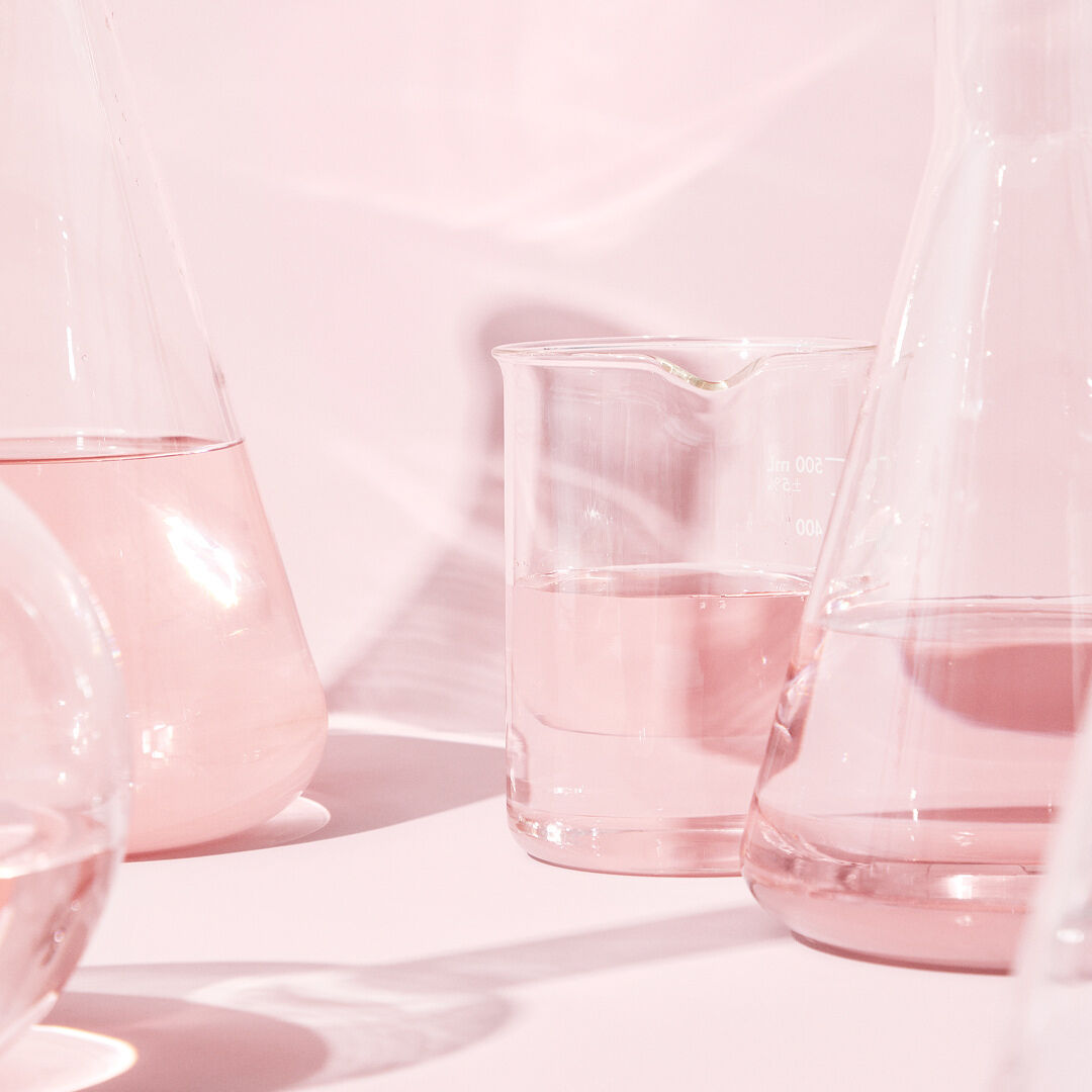 Vasos de precipitados de vidrio transparente con l&iacute;quido incoloro sobre una superficie rosa, reflejando una iluminaci&oacute;n rosada suave.