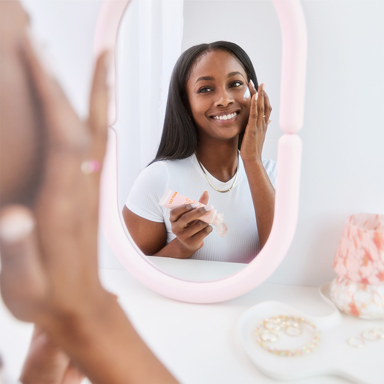 Mujer mirando a un espejo de maquillaje y aplicando el Mary Kay Mineral Facial Sunscreen Broad Spectrum SPF 30 en su mejilla