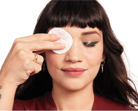 Person gently removing eye makeup with a cotton pad held over one closed eye, demonstrating use of Mary Kay&reg; Oil-Free Eye Makeup Remover. Dark hair, gold jewelry, and a red top are visible against a light background.