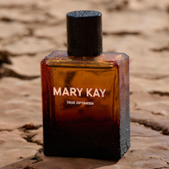 Mary Kay True Optimism fragrance bottle with amber glass and black cap, covered in water droplets and displayed on cracked earth.